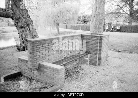 Stone bench in park (1923 - Wethouder Laan - 1939)., Whizgle News, Dutch Desk, The Netherlands, 1950 - 2000. These are the elements in the image. The scene presents a tranquil outdoor setting featuring a rustic brick bench situated near a water body. The bench, built from reddish-brown bricks, has a simple design with a wooden seating surface. Surrounding the bench, lush greenery forms a soft backdrop, consisting of neatly trimmed grass interspersed with patches of white gravel that create a contrasting texture. To one side, a large, gnarled tree with expansive branches adds depth to the scene Stock Photo