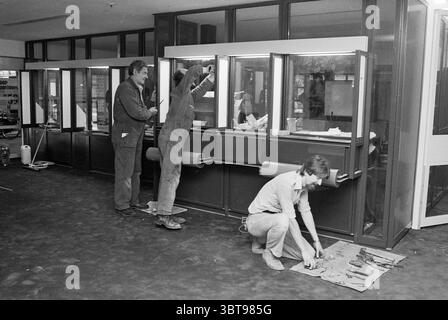 Geesterduin Nutsspaarbank shopping center Castricum, Whizgle News, Dutch Desk, The Netherlands, 1950 - 2000 on 05-1981. The image includes these topics. In a spacious, interior setting that exudes a sense of ongoing work, three individuals are engaged in various tasks within a well-lit area. The floor has a dark, possibly carpeted texture, contrasting sharply with the lighter tones of the room’s walls and windows. Two workers, occupied with the installation or adjustment of a multi-section counter, are positioned near the center. One person, dressed in a dark coverall, stands with a tool in ha Stock Photo