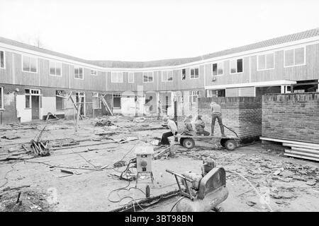 Renovation of homes IJmuiden + Santpoort Renovation IJmuiden The Netherlands, Whizgle News, Dutch Desk, The Netherlands, 1950 - 2000 on 24-12-1980. The image shows these topics. The scene depicts a construction site characterized by a partially built courtyard surrounded by low-rise structures. The buildings have a rustic wooden façade and feature large windows that are yet to be completely installed. The ground is uneven, scattered with construction debris such as bricks, wooden planks, and tools. In the foreground, a group of workers is engaged in various tasks. One individual stands at the Stock Photo