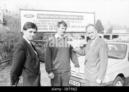Anniversary Mr. Balk HAC Ford dealer Haarlemse Auto central Anniversary anniversaries, Whizgle News, Dutch Desk, The Netherlands, 1950 - 2000 on 02-04-1987. The image contains these topics. The scene unfolds in an outdoor setting, where three men stand together in the foreground. The individuals are dressed in formal attire, with two wearing dark suits and ties, conveying a sense of professionalism, while the third appears in a more casual outfit, likely a button-up shirt or similar. Their expressions range from serious to slightly inquisitive, indicating a potential discussion or interview is Stock Photo