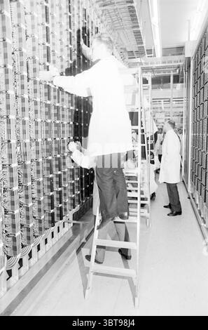 Photo telephone exchange PTT postal services city post, Whizgle News, Dutch Desk, The Netherlands, 1950 - 2000 on 28-04-1967. These are the elements in the image. The setting appears to be a large, industrial space filled with rows of intricate machinery and electronic equipment. The architecture suggests a technological environment, likely a laboratory or a data processing facility. Several individuals in white lab coats are busy at work, indicating a professional atmosphere. One person is positioned on a ladder, reaching up to connect or adjust components in the machinery, while another indi Stock Photo