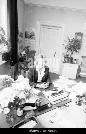 Information officer Jeanine van Ooyen Zandvoort Zandvoort Zandvoort, Whizgle News, Dutch Desk, The Netherlands, 1950 - 2000 on 08-06-1989. The image shows these topics. The scene is set in an office, characterized by a neat and organized atmosphere. A woman sits confidently at a wooden desk, her bright smile adding warmth to the space. She is dressed in a pinstriped suit, creating a professional appearance. The desk is cluttered but managed, featuring several office supplies such as typewriters and a pen holder filled with various pens and pencils. Next to these tools, there is a partially fil Stock Photo