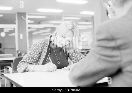 Monique van der Ven takes driving test C.B.R., Whizgle News, Dutch Desk, The Netherlands, 1950 - 2000 on 07-03-1974. The image shows these topics. In a well-lit indoor setting, a young woman sits at a table, focused intently on her task as she writes. She has shoulder-length blonde hair styled in soft waves that frame her face. Dressed in a patterned blouse with floral designs, she adds a touch of vibrancy to the otherwise neutral environment. The table surface is simple and unadorned, emphasizing her concentration on her work. In the background, a blurred figure can be seen, possibly another Stock Photo