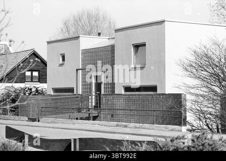 Dike house Kerkstraat. 55 Haarlem Kerkstraat The Netherlands, Whizgle News, Dutch Desk, The Netherlands, 1950 - 2000 on 26-03-1991. These are the elements in the image. The scene depicts a modern architectural structure characterized by clean lines and a minimalist design. The building features two prominent cubes with a smooth, white exterior, creating a striking contrast against the textured, dark brown shingles of a traditional house situated to the left. In front of the modern building, a gently sloping pathway leads up to a visible entrance, emphasizing accessibility. A large glass block Stock Photo