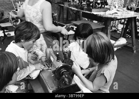 Stuif Stuif children at a sewing machine, Whizgle News, Dutch Desk, The Netherlands, 1950 - 2000 on 16-08-1979. The image includes these topics. The scene captures a bustling workshop filled with activity and familial warmth. In the foreground, two children are engaged in sewing, their focused expressions displaying a mix of concentration and curiosity. One child, with short hair, leans forward, examining the sewing machine's needle, while the other, with slightly longer hair, deftly guides fabric through the machine. The metallic surface of the sewing machine glints in the available light. Ne Stock Photo
