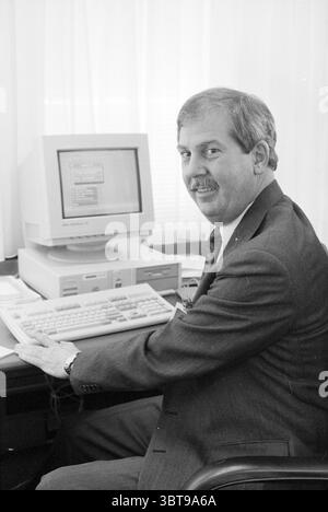 Mr. Leadridge Fluor Daniel, Whizgle News, Dutch Desk, The Netherlands, 1950 - 2000 on 03-08-1994. The image includes these topics. The scene features a man seated at a desk, exuding a professional demeanor. He is dressed in a tailored dark suit with a collared shirt. His neatly combed hair and mustache contribute to a polished appearance. The man is turning slightly towards the viewer, wearing a warm, confident smile, indicating a friendly and approachable attitude. In front of him, an older-style computer with a bulky monitor and a keyboard dominates the workspace. The monitor displays a simp Stock Photo