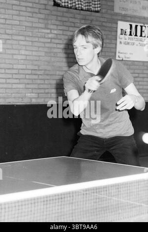 Table tennis player of TTNV (Table Tennis Association Nieuw-Vennep), Whizgle News, Dutch Desk, The Netherlands, 1950 - 2000 on 11-11-1984. The image contains these topics. In a dimly lit indoor setting, a young person is actively engaged in a game of table tennis. They have short, light-colored hair and are focused intently on the ball, demonstrating concentration and athleticism. The player is wearing a collared shirt, likely in a muted color, paired with dark trousers, reflecting a casual yet sporty appearance. The table tennis equipment is prominent in the scene, with a sleek, rectangular t Stock Photo