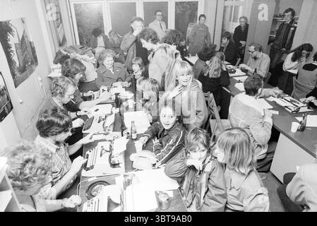 Making Sinterklaas poems Haarlems Dagblad St. Nicholas Sinterklaas, Whizgle News, Dutch Desk, The Netherlands, 1950 - 2000 on 24-11-1978. The image shows these topics. The scene is bustling and dynamic, depicting a gathering in a cluttered, warmly lit room filled with a mix of aged wooden furniture and retro decor. Numerous people, ranging from children to adults, are engaged in various activities, creating a lively atmosphere. In the foreground, several tables are strewn with typewriters and sheets of paper, indicating a collaborative effort, perhaps in writing or journalism. A group of indiv Stock Photo