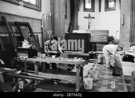 Restoration of organ in church.., Whizgle News, Dutch Desk, The Netherlands, 1950 - 2000. These topics are shown in the image. The scene depicts a spacious interior, likely a church or an old community building, characterized by tall ceilings and stone walls, lending it an air of historical significance. The natural light filtering through stained glass windows casts colorful patterns on the floor, creating a vibrant contrast against the muted tones of the surroundings. In the foreground, two individuals are engaged in carpentry work. One person, dressed in a plaid shirt, is seated at a sturdy Stock Photo