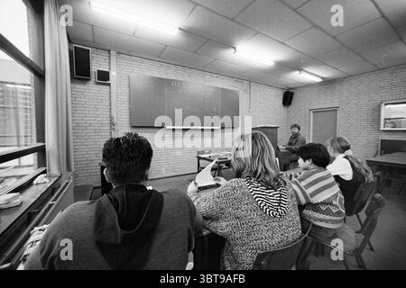 Class photo Delta School, Whizgle News, Dutch Desk, The Netherlands, 1950 - 2000 on 12-01-1993. These topics appear in the image. The scene features a classroom setting characterized by a casual yet focused atmosphere. The room has light-colored brick walls and a ceiling with rectangular tiles, creating a clean, functional space. The lighting is soft and even, likely from fluorescent fixtures, providing a bright environment for learning. In the foreground, three individuals are seated at desks, their backs to the viewer. The first person on the left has short, dark hair and is dressed in a dar Stock Photo