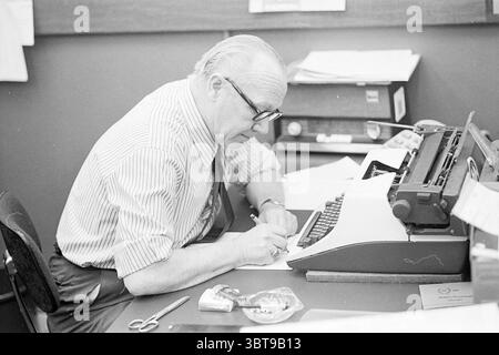 Jan van Dam during work Editorial IJmuider Courant Personen newspapers and magazines, Whizgle News, Dutch Desk, The Netherlands, 1950 - 2000 on 23-03-1976. These topics appear in the image. In a cozy office setting, an elderly man sits at a wooden desk, deeply focused on his work. He is wearing a white and gray striped dress shirt with rolled sleeves, complemented by a dark tie. His glasses sit low on his nose, and his thinning hair is neatly combed back. The desk is slightly cluttered, with scattered sheets of paper, a pair of scissors resting to the side, and an open typewriter that dominate Stock Photo