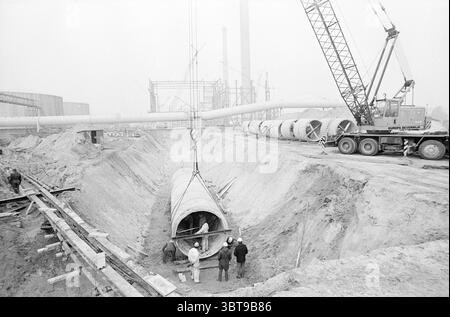 Laying pipes Velsen. Work, Whizgle News, Dutch Desk, The Netherlands, 1950 - 2000 on 24-02-1972. The image includes these topics. The scene depicts a construction site, characterized by a sense of industrious activity and a muted color palette. The ground is a mix of dirt and sand, suggesting recent excavation, with large trenches cutting through the site. In the foreground, several workers are engaged in their tasks near a large concrete pipe that lies on its side, partially obscured by the trench. Their clothing is practical—overalls and hard hats, reflecting a focus on safety amid the rough Stock Photo