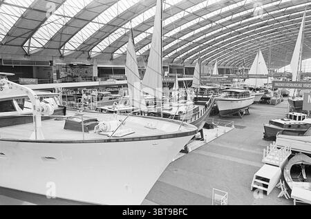 Hiswa exhibition R.A.I. Exhibitions, Whizgle News, Dutch Desk, The Netherlands, 1950 - 2000 on 16-03-1964. The image shows these topics. The scene features an expansive indoor marina, characterized by a high, vaulted glass ceiling that allows natural light to pour in, illuminating the space. The architecture is modern, with sleek lines and a spacious layout. In the foreground, various yachts and sailboats are meticulously arranged, showcasing different styles and sizes. The hulls of the boats gleam in shades of white and navy, reflecting the overhead light. Ropes and rigging are neatly coiled, Stock Photo