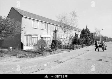 Rental properties Hondsdrafstraat Nieuw-Vennep Houses and house construction Nieuw-Vennep Hondsdrafstraat, Whizgle News, Dutch Desk, The Netherlands, 1950 - 2000 on 21-04-1987. These are the topics in the image. The scene depicts a residential street with a row of modest, two-story houses lined up in a neat formation. The architectural style is simple, with rectangular shapes and a sloped roof, all rendered in muted tones of beige and gray. The facade features large windows adorned with sheer curtains, giving a glimpse of the indoors while maintaining a sense of privacy. In the foreground, a c Stock Photo