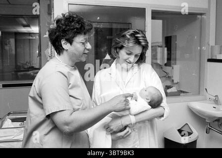 Pediatrician Dahlberg with baby, Whizgle News, Dutch Desk, The Netherlands, 1950 - 2000 on 11-05-1990. The image includes these topics. In a softly lit hospital room, two women are engaged in a tender moment with a newborn baby. The walls are a muted pastel, contributing to a calming atmosphere. The first woman, wearing light blue scrubs, has curly hair and glasses. Her expression is warm and inviting as she looks down at the baby. She is gently pointing towards the infant, perhaps offering encouragement or sharing a moment of connection. The second woman, dressed in a white lab coat over a li Stock Photo