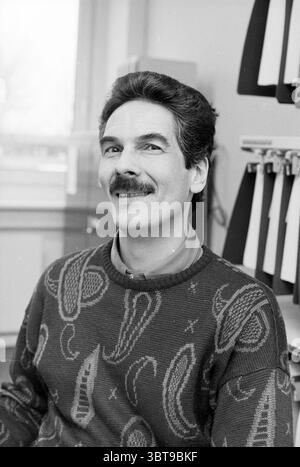 Peter Abrahams Media Systems, Whizgle News, Dutch Desk, The Netherlands, 1950 - 2000 on 27-11-1990. The image includes these topics. The scene features a man with medium-length dark hair, styled in a somewhat voluminous manner, and a prominent, well-groomed mustache. He has a warm, inviting smile that conveys a sense of friendliness and approachability. He is seated at a desk, and the backdrop includes a series of folders or files neatly organized, likely suggesting a professional or office environment. The colors in the setting are muted, with shades of gray dominating the scene, which adds a Stock Photo