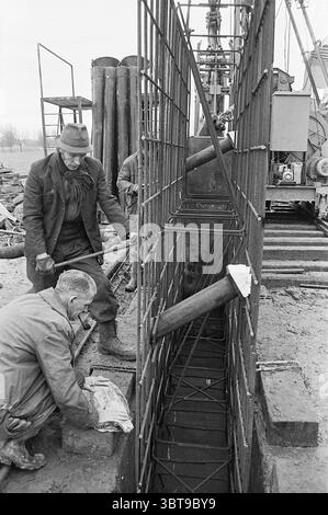 Building with reinforced concrete, Whizgle News, Dutch Desk, The Netherlands, 1950 - 2000 on 04-1970. The image contains these topics. The scene captures a construction site, characterized by a gritty, industrial atmosphere. In the foreground, two workers are engaged in an activity that suggests they are setting up or checking equipment. One worker, kneeling, appears focused on a task with a piece of cloth in his hand, seemingly inspecting or cleaning something. His clothing is somewhat worn, predominantly dark, adding to the rugged aesthetic of the setting. The other worker stands slightly to Stock Photo