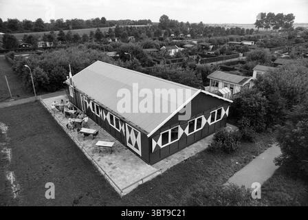 Clubhouse Poelpolder, Whizgle News, Dutch Desk, The Netherlands, 1950 - 2000 on 11-08-1991. The image includes these topics. The scene features a large wooden structure with a slanted roof, painted in dark hues with contrasting white diamond patterns on its facade. Surrounding the building is a spacious outdoor area, equipped with several tables and chairs, likely intended for gatherings or leisure activities. The setting is characterized by lush greenery, with a variety of trees, shrubs, and low vegetation surrounding the structure. In the background, a series of smaller buildings, likely cab Stock Photo