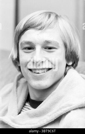 Portrait of a man., Whizgle News, Dutch Desk, The Netherlands, 1950 - 2000. These topics appear in the image. The scene features a young individual with a warm and inviting smile, exuding an approachable demeanor. He has short, straight blonde hair that curls slightly at the ends, framing his face. His complexion is fair, showcasing a scattering of light freckles across his cheeks. He is dressed in a light-colored, plush garment that appears soft and comfortable, adding to the relaxed atmosphere of the moment. Beneath the outer layer, subtle horizontal stripes in darker tones are visible, hint Stock Photo