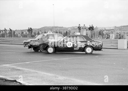 Crazy Race Zandvoort Circuit Zandvoort Zandvoort, Whizgle News, Dutch Desk, The Netherlands, 1950 - 2000 on 26-12-1984. These are the topics in the image. The scene depicts a dramatic moment during a car race, showcasing two cars in an intense collision. One vehicle, painted in matte black, bears the number “27” prominently on its side. The other car, in stark contrast, is a lighter shade, possibly silver or white, and marked with the number “71.” The black car exhibits significant damage to its front end, crumpled and battered, while the lighter car has a crushed rear section, hinting at the Stock Photo