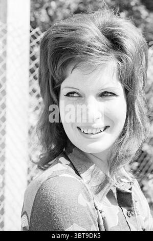 Models Loe-Loe Models, Whizgle News, Dutch Desk, The Netherlands, 1950 - 2000 on 29-05-1971. The image shows these topics. The scene features a woman with striking, voluminous hair styled with a distinct 1960s flair, characterized by a rich auburn hue. Her expression radiates warmth and confidence, highlighted by a bright, engaging smile that reveals her cheerful demeanor. She has bold eyeliner accentuating her eyes, adding to her expressive features. The background suggests an outdoor setting, with soft, dappled sunlight filtering through lush foliage. Behind her, a chain-link fence is visibl Stock Photo
