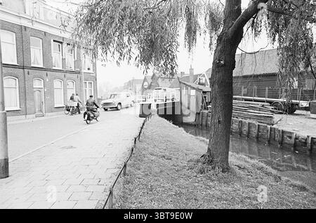 Too narrow bridge on the Schalkwijkerstraatweg Traffic safe traffic Haarlem Schalkwijkerstraat The Netherlands, Whizgle News, Dutch Desk, The Netherlands, 1950 - 2000 on 26-10-1964. These are the topics in the image. The scene depicts a quaint, town-like setting, captured in monochrome tones that evoke a sense of nostalgia. On one side of a narrow, cobblestone street, a gentle slope leads down to a tranquil canal bordered by lush grass. A large tree stands prominently, its branches extending gracefully, casting an intricate pattern of shadows on the walkway. Across the street, a row of classic Stock Photo