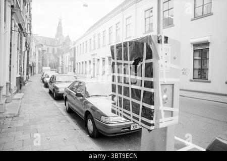 Packed parking meters, Whizgle News, Dutch Desk, The Netherlands, 1950 - 2000 on 31-10-1991. These topics are shown in the image. The scene unfolds in a narrow urban street lined with calm residential buildings. The architecture features a mix of traditional and contemporary designs, with whitewashed facades that are simple yet inviting. Along the sidewalk, a series of parked cars—mostly in muted tones of gray and silver—are neatly aligned, evoking a sense of order. To one side, a utility box stands out, caught in the act of being obscured by a patchy arrangement of black tape and grid pattern Stock Photo