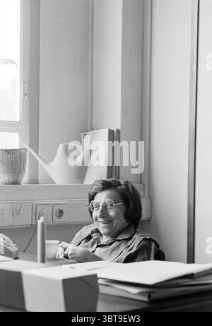 Portrait of Dr Lea Dasberg University of Amsterdam Portraits Amsterdam The Netherlands, Whizgle News, Dutch Desk, The Netherlands, 1950 - 2000 on 11-06-1980. The image shows these topics. In a well-lit room, an older woman sits at a desk, her demeanor warm and inviting. She has short, dark hair and wears large, round glasses that accentuate her expressive eyes. Her lips curl into a smile, suggesting a sense of joy or amusement. The setting features a mix of bright and muted colors. The background is adorned with soft beige walls, contributing to a calm atmosphere. Natural light streams in from Stock Photo