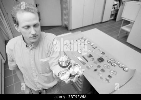 Seizure[name] car markings Hoofddorp The Netherlands, Whizgle News, Dutch Desk, The Netherlands, 1950 - 2000 on 26-03-1992. These are the topics in the image. In a well-lit indoor setting, the scene captures a man attentively examining a collection of circular emblems, possibly car badges. The man is dressed in a light-colored, button-up shirt, and he appears focused, holding one of the badges in his right hand while looking at it, suggesting a sense of curiosity or nostalgia. The background features a worktable covered with a smooth, neutral-toned surface, on which numerous similar badges are Stock Photo
