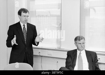 Northern Telecom press conference, Whizgle News, Dutch Desk, The Netherlands, 1950 - 2000 on 11-05-1993. These are the elements in the image. In a bright, modern room with large windows allowing natural light to flood in, two men are engaged in a professional setting. The man standing on the left has a distinguished air about him, dressed in a dark suit paired with a patterned tie. His hand gestures emphasize his points, conveying a sense of authority and engagement. He appears expressive, his face focused and slightly animated, suggesting he is addressing an important topic. Seated to his rig Stock Photo
