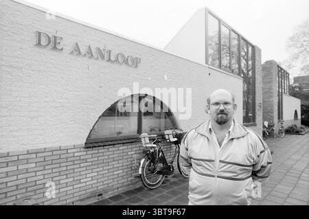 Man at the Aanloop sports center Kruisweg Hoofddorp Kruisweg The Netherlands, Whizgle News, Dutch Desk, The Netherlands, 1950 - 2000 on 12-04-1989. These topics are shown in the image. The scene captures a man standing in front of a modern building characterized by a blend of smooth and textured surfaces. The architecture features a prominent arch shape integrated into a brick wall, contrasting with the sleek, angular lines of glass and light-colored materials above. The man is dressed in a light-colored, zippered jacket, which complements the muted tones of the surroundings. His expression is Stock Photo