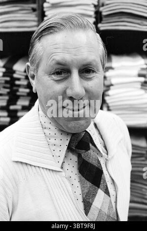 Gentleman in front of cupboard with folded shirts in clothing store, Whizgle News, Dutch Desk, The Netherlands, 1950 - 2000 on 02-1982. The image contains these topics. A well-dressed man stands confidently, offering a warm smile that conveys friendliness and approachability. His hair is neatly styled, showcasing a blend of silver and light tones, contributing to an air of wisdom. He wears a light colored, textured sweater over a collared shirt that features a pattern of dots, adding a touch of personality. The tie is bold with a mix of checkered patterns in earthy tones, drawing attention to Stock Photo