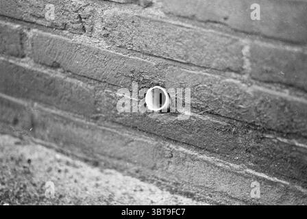 Mrs van Gool of the house in poor condition Houses and house construction, Whizgle News, Dutch Desk, The Netherlands, 1950 - 2000 on 18-01-1983. These topics appear in the image. The scene presents a close-up view of a brick wall, characterized by a slightly rough texture, with the bricks displaying varying shades of gray. Some bricks show subtle signs of wear, with small cracks and uneven surfaces adding to the character of the wall. A cylindrical hole, likely a pipe, protrudes from the wall, appearing slightly weathered, with edges that are smooth yet worn. The color palette is dominated by Stock Photo