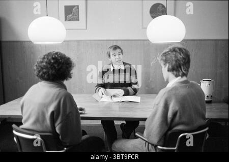 Mr. Jongbloed RSAD Haarlem Persons Haarlem The Netherlands, Whizgle News, Dutch Desk, The Netherlands, 1950 - 2000 on 08-01-1985. These topics are shown in the image. In a cozy, warmly lit setting, three individuals are gathered around a wooden table. The warm tones of the wood panels on the walls create an inviting atmosphere, complemented by the soft glow of two round pendant lights hanging from the ceiling, casting gentle illumination over the scene. At the center of attention is a man with short, light brown hair, dressed in a striped sweater featuring various shades of blue and brown. He Stock Photo