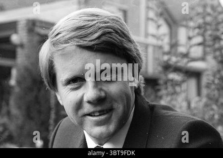 Portrait Jos Draayer Hillegom Portraits Hillegom, Whizgle News, Dutch Desk, The Netherlands, 1950 - 2000 on 16-09-1982. These topics appear in the image. In the scene, a young man is the focal point, exuding a warm and approachable demeanor. He has neatly styled hair that appears light in color, possibly a shade of blonde or light brown, creating a contrast against the darker, formal attire he wears. His jacket is tailored and dark, typically associated with business or formal settings. The background suggests an outdoor setting with blurred, indistinct elements hinting at greenery and structu Stock Photo