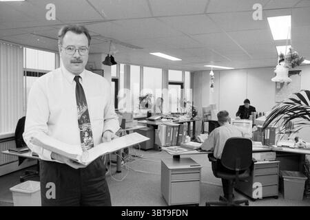 Willem Verhoog PTT Leiden Leiden, Whizgle News, Dutch Desk, The Netherlands, 1950 - 2000 on 17-12-1996. These are the topics in the image. The scene shows a spacious office environment, characterized by a mix of workstations and large windows that allow natural light to flood in, creating a bright and open atmosphere. In the foreground, a middle-aged man stands holding an open binder, his expression appears focused and approachable. He wears a white dress shirt complemented by a colorful tie, which features a festive theme—likely suggesting a seasonal celebration. His glasses rest just below h Stock Photo