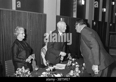 Jub. mr. Otte Jubilee anniversaries, Whizgle News, Dutch Desk, The Netherlands, 1950 - 2000 on 18-08-1972. The image shows these topics. The scene showcases a formal indoor setting, characterized by its muted, dark wooden paneling that creates a warm, inviting atmosphere. A long table is prominent, adorned with modest floral arrangements that add a touch of elegance. At the table, a man in a dark suit stands, engaging warmly with another man, who is leaning slightly forward, seemingly in conversation or greeting. To one side, a woman wearing a black dress and glasses watches the interaction wi Stock Photo