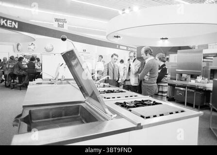 Horecava RAI Amsterdam Horeca, Whizgle News, Dutch Desk, The Netherlands, 1950 - 2000 on 12-01-1977. These are the topics in the image. The scene is set in a modern kitchen exhibition, showcasing various cooking and kitchen appliances. The foreground features a large, sleek kitchen island with a deep sink and multiple burners arranged neatly. The appliances are metallic and polished, reflecting the overhead lights. A small group of people is gathered around the island; their attention is drawn to a demonstration of the kitchen setup. There are two men and a woman in business attire, engaged in Stock Photo