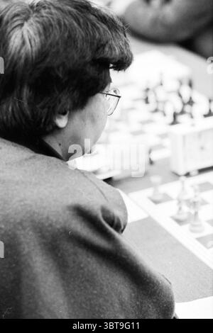 Blastovens Chess Tournament Wijk aan Zee, Whizgle News, Dutch Desk, The Netherlands, 1950 - 2000 on 28-01-1991. These are the elements in the image. In the scene, a person is seated at a chess table, their back facing the viewer. The individual has medium-length, dark hair and is wearing glasses, which catch a hint of light. They are draped in a black garment, possibly a coat or sweater, which adds a somber tone to the setting. The chessboard in front of them features a classic checkered pattern, where the pieces are arranged in mid-game. The pieces themselves are intricately shaped, contrasti Stock Photo