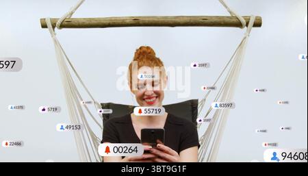Smiling red-haired woman holding phone in rope wooden hammock chair at studio with notifications Stock Photo