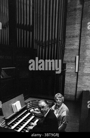 Organizers Bartelink in basilica with organ Organs and organists, Whizgle News, Dutch Desk, The Netherlands, 1950 - 2000 on 12-10-1988. The image shows these topics. In a dimly lit space, two individuals are seated near an elaborately designed organ. The organ features numerous keys and various control buttons, showcasing a rich, polished wood surface that contrasts with the more industrial, textured brick walls behind it. The scene is dominated by towering pipes that rise majestically, adding a sense of grandeur and depth to the setting. To the left, an older man, wearing spectacles, is focus Stock Photo