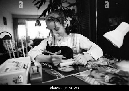 Monique Barneveld Big Dad, Whizgle News, Dutch Desk, The Netherlands, 1950 - 2000 on 22-01-1991. These are the elements in the image. In a cozy interior setting, a young woman sits at a table, her expression focused and content. She has short, slightly tousled hair and wears a light-colored long-sleeve shirt paired with denim overalls. Resting on her lap is a small black rabbit, its ears perked up, creating a sense of calmness in the scene. The table is cluttered with an array of magazines, vividly arranged with their covers facing up, showcasing colorful photographs and bold designs. In front Stock Photo