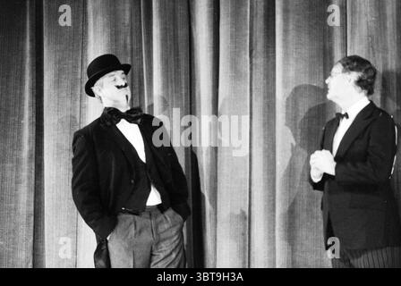 Heerenleed theatre, Whizgle News, Dutch Desk, The Netherlands, 1950 - 2000 on 15-10-1990. These topics appear in the image. The scene depicts two men performing on a stage, with a rich, textured curtain in the background that drapes elegantly, likely in a dark hue. One man stands confidently, adopting a theatrical pose, dressed in a classic black tuxedo with a top hat, his face adorned with a comical moustache that adds a playful character to his appearance. His stance, slightly tilted, suggests a sense of amusement or bravado. The second man, wearing spectacles, is also attired in formal wear Stock Photo