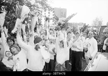 Four-day evening with cheering children., Whizgle News, Dutch Desk, The Netherlands, 1950 - 2000. These are the topics in the image. The scene captures a lively, celebratory atmosphere with a large group of people gathered together outdoors. Many individuals are holding vibrant bouquets, possibly of flowers or leafy branches, raised high above their heads as they express excitement. In the foreground, a collection of children stands out; their youthful faces are filled with joy and eagerness. They wear simple, light-colored clothing, perhaps sweatshirts, creating a sense of unity among them. T Stock Photo