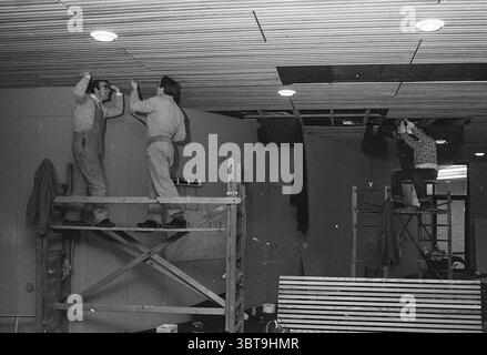 Setting up an office., Whizgle News, Dutch Desk, The Netherlands, 1950 - 2000. The image contains these topics. The scene captures a dynamic moment within an industrial or commercial setting undergoing renovation or construction. **People**: Three individuals are engaged in activities related to overhead work. Two men are positioned on wooden scaffolding, likely carefully installing or adjusting the ceiling panels. Both men wear practical work attire, including shirts and overalls, which provide a sturdy appearance conducive to labor. The man on the left has his hands raised, possibly holding Stock Photo