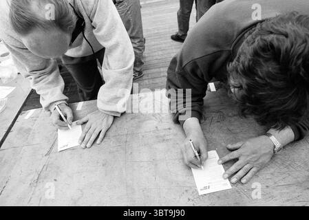 Register with the FNV Velsen strike fund Velsen, Whizgle News, Dutch Desk, The Netherlands, 1950 - 2000 on 14-04-1991. These are the topics in the image. In this scene, two individuals are bent over a wooden table, deeply focused on their task. Each person is positioned closely, their heads nearly touching as they write. The surface of the table exhibits a weathered texture, marked with scratches and a natural patina from frequent use. The person on the left wears a light gray sweatshirt, with their hand gripping a pen, poised over a blank card that rests on the table. They appear absorbed in Stock Photo