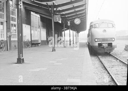 Platform station IJm. Station, Whizgle News, Dutch Desk, The Netherlands, 1950 - 2000 on 10-05-1977. The image shows these topics. The scene features a train station platform characterized by its industrial charm and a nostalgic atmosphere. The architecture is marked by a long, overhanging roof that provides shelter, supported by wooden beams painted in dark tones. The flooring comprises large, rectangular paving stones, which create a rhythmic pattern leading towards the train tracks. To the right, a vintage train stands idly, painted in muted tones of gray. Its rounded front and distinct des Stock Photo