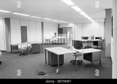 Workspace with multiple desks in new office, Whizgle News, Dutch Desk, The Netherlands, 1950 - 2000 on 19-06-1978. These topics appear in the image. The scene depicts a spacious, empty office environment characterized by minimalistic decor and a monochromatic palette. The flooring is covered in dark carpeting that contrasts with the light-colored walls, which are predominantly white with a few sections featuring a muted gray. Several desks are arranged throughout the room, each comprising a simple, modern design with clean lines. The desks are furnished with metal frames and flat surfaces, som Stock Photo