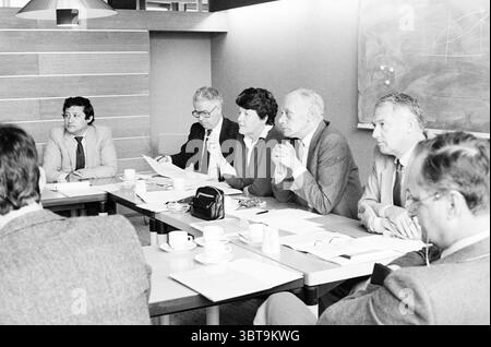 Press conference IJmond municipalities Press conference, Whizgle News, Dutch Desk, The Netherlands, 1950 - 2000 on 09-05-1983. These topics are shown in the image. In a conference room, a group of six individuals is gathered around a long, rectangular table made of light-colored wood. The setting has a professional atmosphere, characterized by the muted tones of the furnishings and wall. The individuals are dressed in formal attire, with a mix of light and dark suits. Their expressions convey focus and seriousness, as they lean slightly forward, engaged in discussion. A man on the far left has Stock Photo