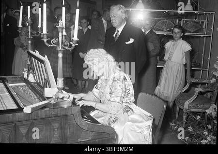 Piano recital., Whizgle News, Dutch Desk, The Netherlands, 1950 - 2000. The image includes these topics. The scene captures a vintage gathering in a well-decorated room that exudes elegance and a subtle air of nostalgia. At the forefront, a woman sits gracefully at a grand piano, her long, flowing dress shimmering with intricate patterns and soft hues that suggest both refinement and drama. Her hair is styled in soft curls, perhaps adorned with a decorative piece that complements her attire, drawing attention to her focused expression as she plays. Surrounding her are several individuals, some Stock Photo